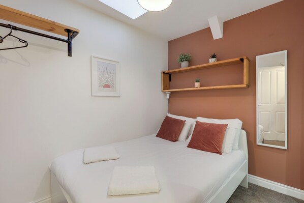 Un lit bien fait, avec du linge de lit blanc et deux oreillers marron, une étagère en bois ornée de plantes, un tableau encadré accroché au mur et un miroir qui reflète la porte.