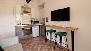 Cocina compacta con armarios blancos, microondas, refrigerador pequeño y barra de madera con dos taburetes verdes.