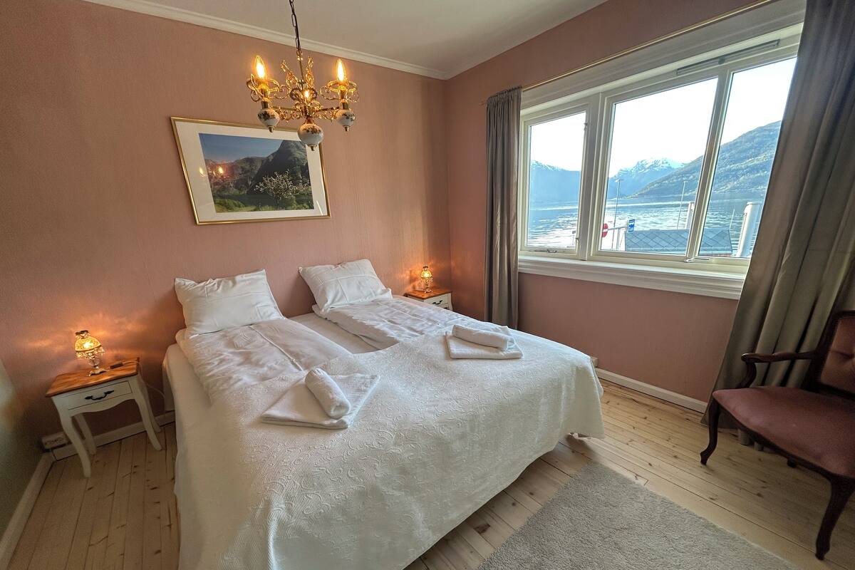 A bedroom with a large bed, two bedside tables with lamps, a chandelier, a window with a view of the sea and mountains, and a chair.