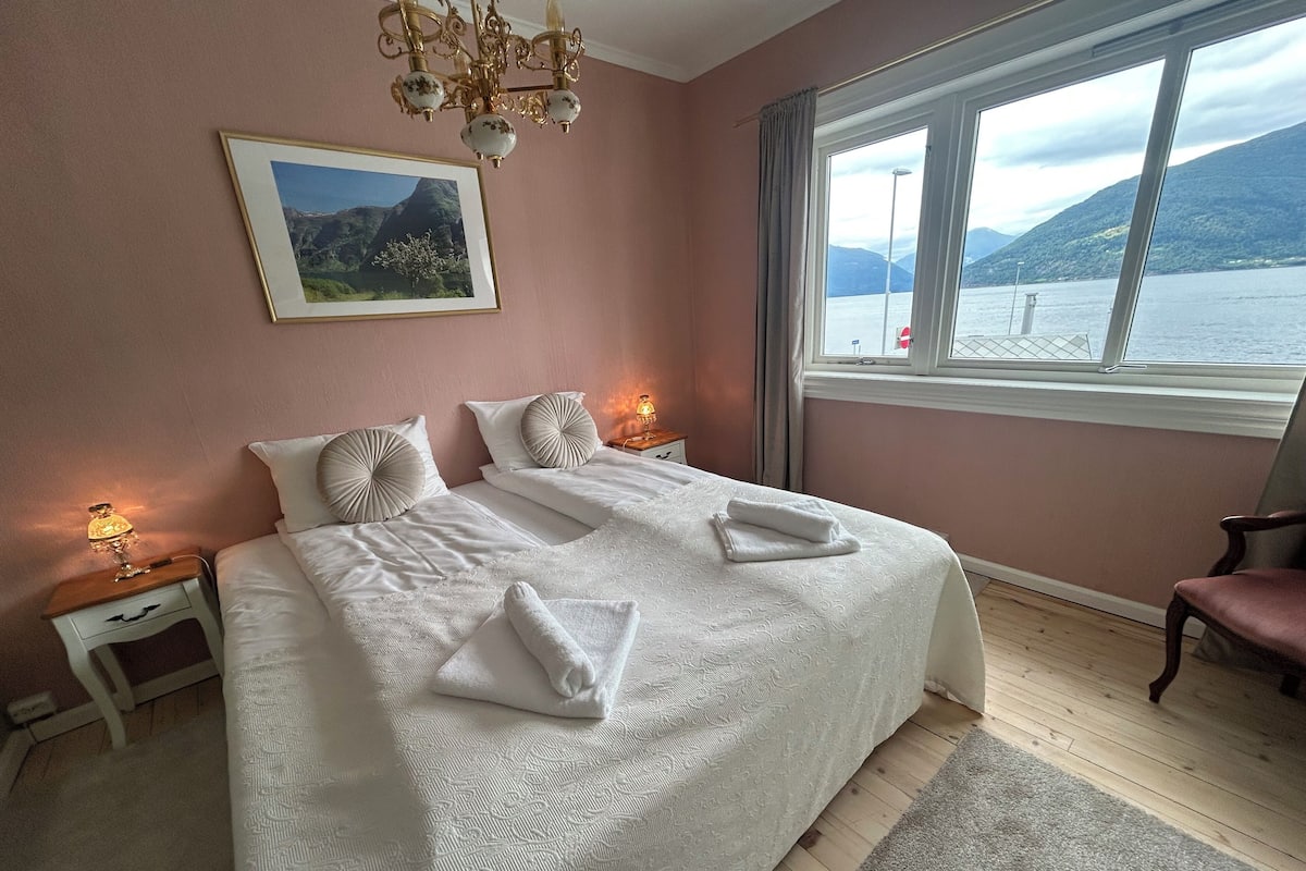 A bedroom with a bed, two bedside tables, a chandelier, a window with a view of mountains, and a framed picture on the wall.