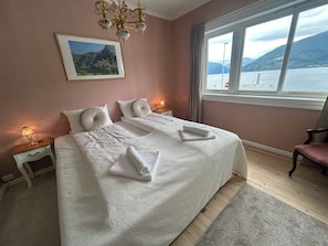 Une chambre à coucher avec un lit, deux tables de chevet, un lustre, une fenêtre donnant sur les montagnes et un tableau encadré au mur.