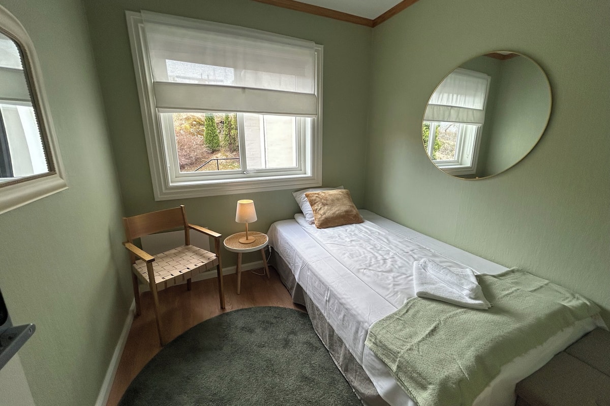 A small, single-bed room with a chair, a small table, a round mirror, and a window with a view of greenery outside.
