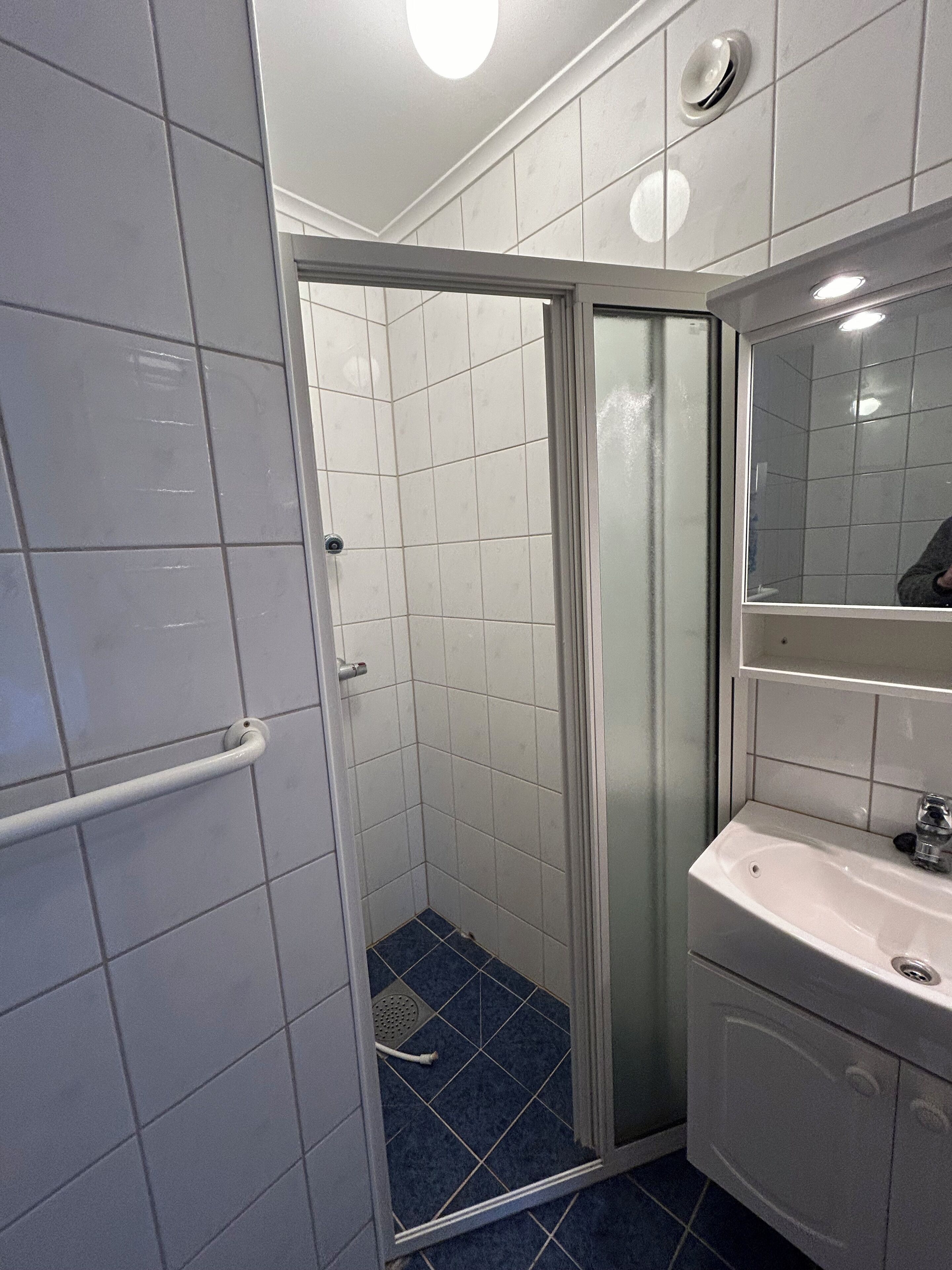 Un baño con paredes de azulejos blancos, mampara de ducha de vidrio, piso de azulejos azules y un lavamanos blanco con espejo arriba.