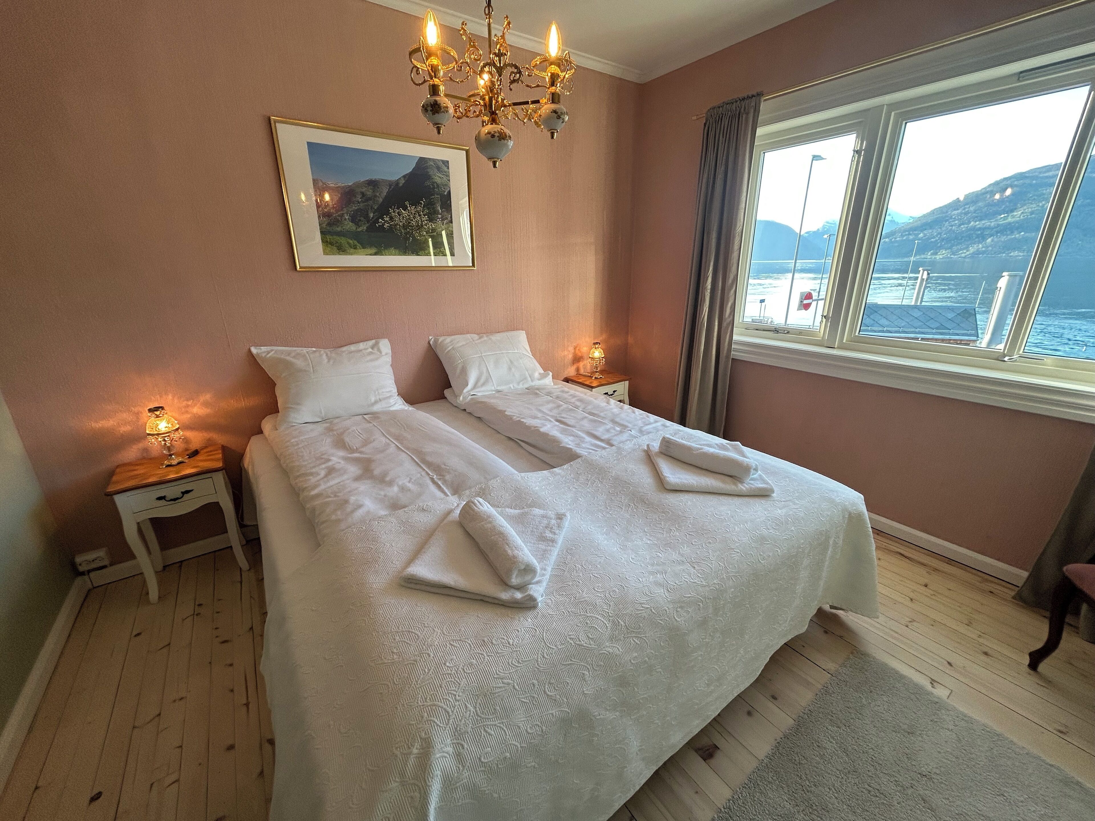 A neatly made bed with white linens and towels, a chandelier, a framed picture on the wall, and a window with a view of the sea.