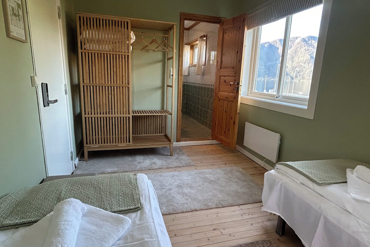 A bedroom with two beds, a wooden wardrobe, a window with a mountain view, and a door leading to a bathroom.