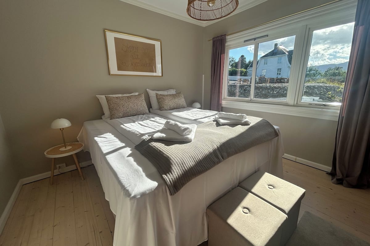 A bedroom with a large bed, a bedside table, a lamp, a window with curtains, and a framed picture on the wall.