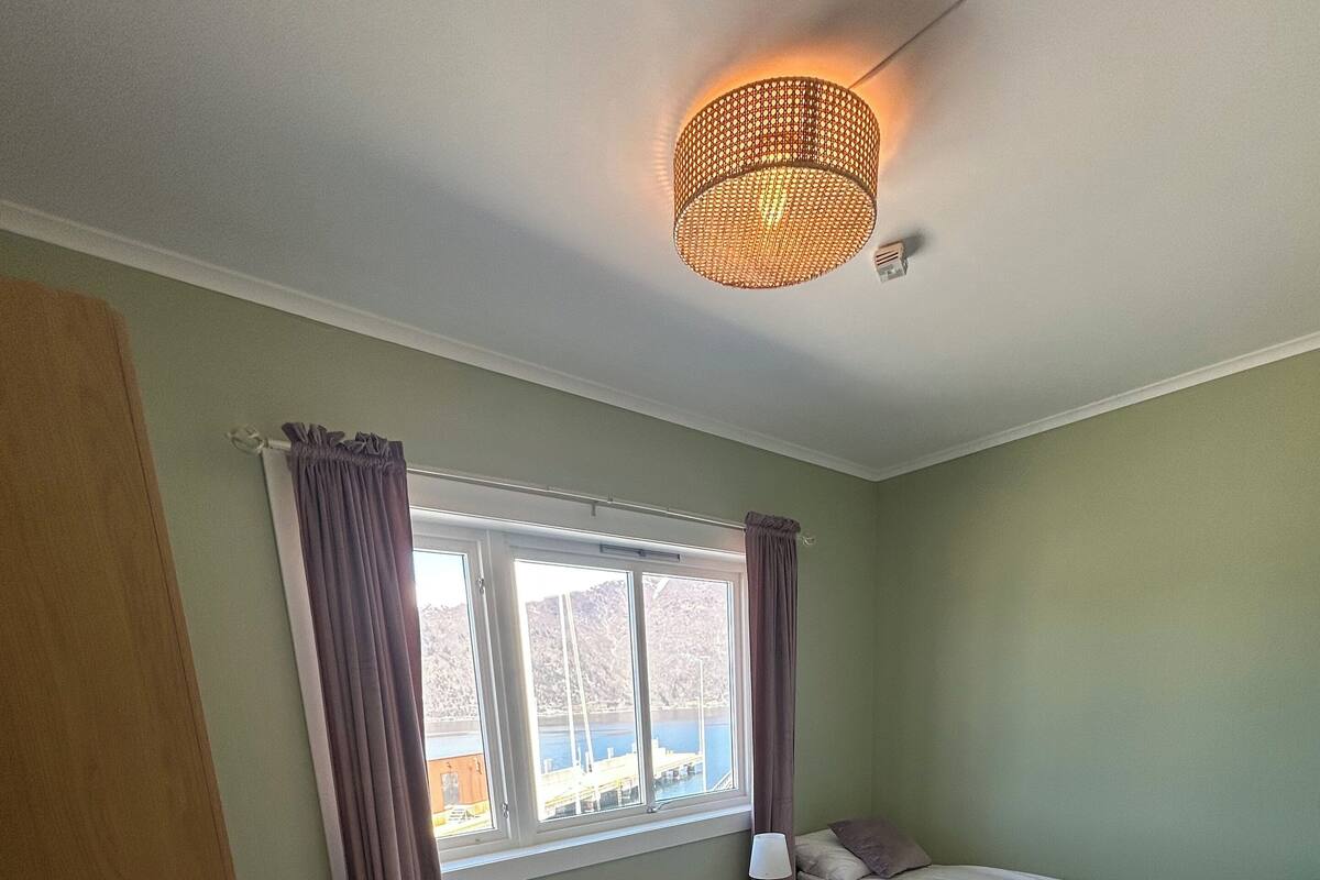 A bedroom with a bed, a bedside table, a lamp, a window with curtains, and a ceiling light.