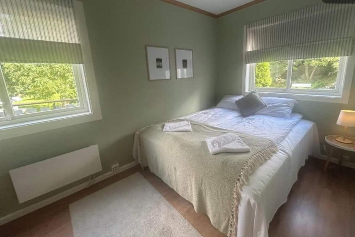 A bedroom with a bed, two framed pictures, a bedside lamp, and a window with blinds.