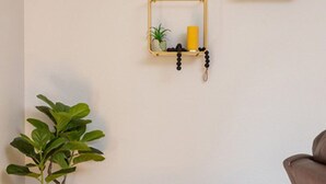 Una esquina de una habitación con suelo de madera, pared blanca, una planta pequeña en maceta, un taburete redondo tejido y una lámpara de pie con pantalla estampada.