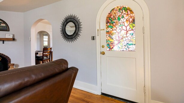 Una puerta blanca con un vitral, un espejo circular negro y un banco de cuero marrón en una habitación con suelo de madera y paredes blancas.