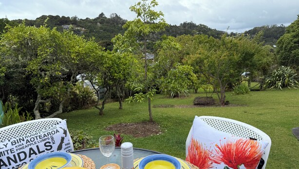 A table set outdoors with fruits, a bottle, and a glass, surrounded by greenery and trees. - Retro Russell Retreat - Mins to Ferry, Beach, Eats (Russell)