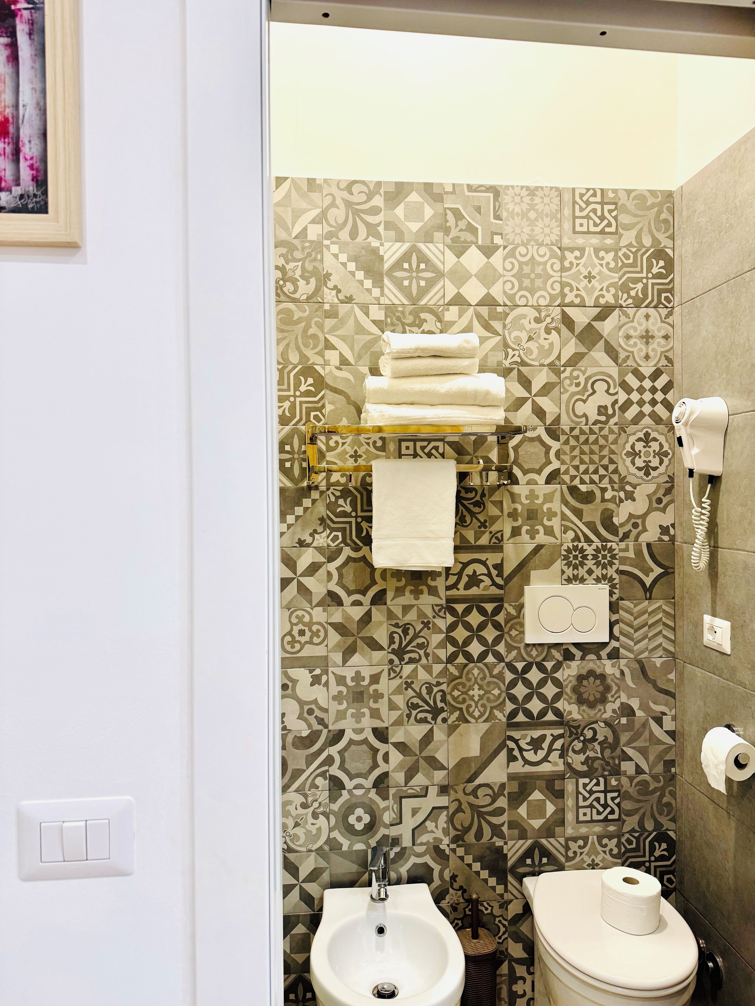 Ein Badezimmer mit gemusterten Fliesen, einem Bidet, einer Toilette mit Toilettenpapierhalter und einem Handtuchhalter mit gefalteten Handtüchern.