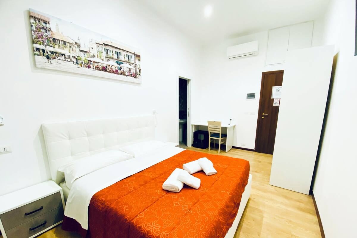 A hotel room with a large bed, a desk, and a chair. There is a painting on the wall and an air conditioning unit.