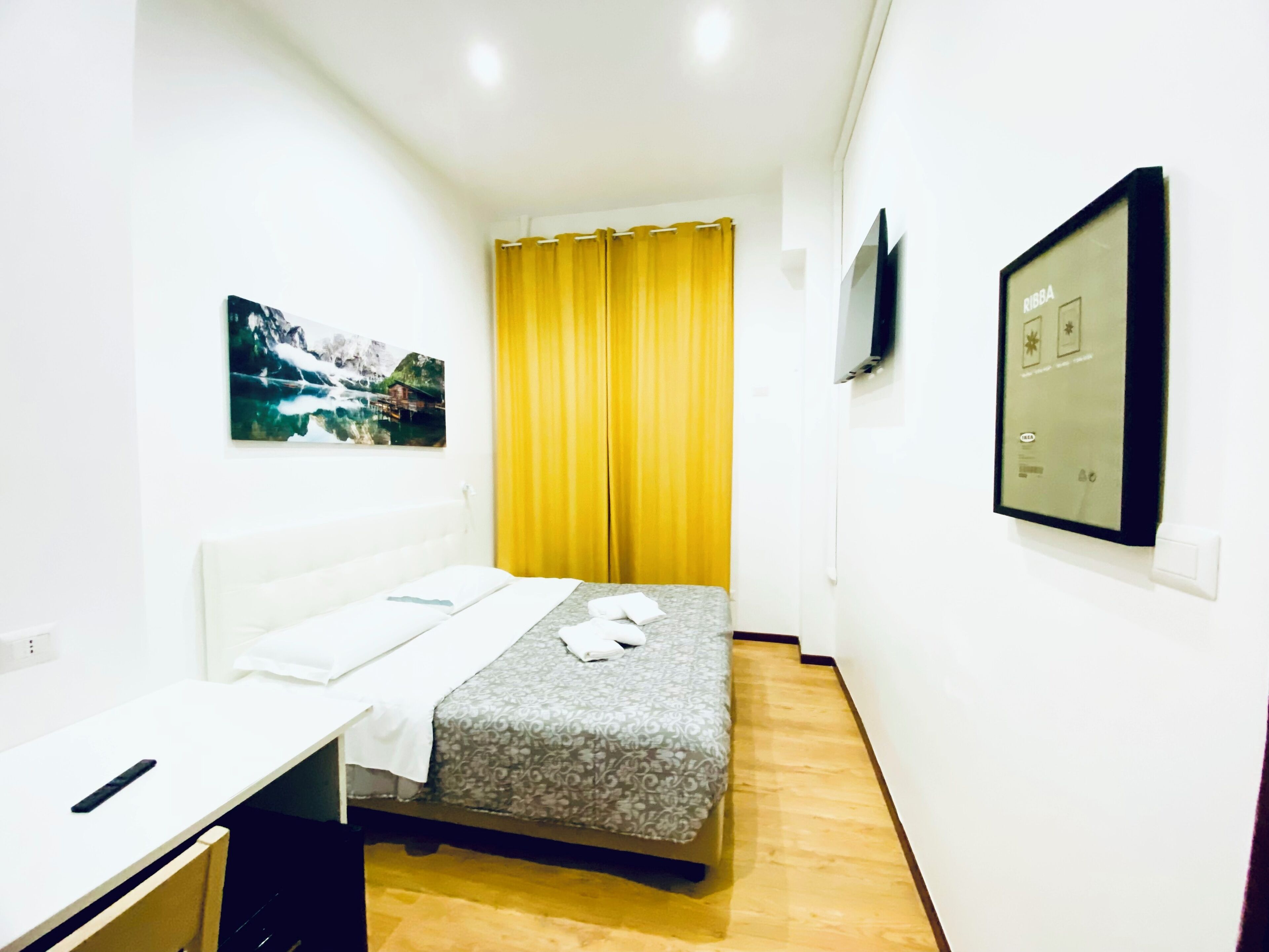 A hotel room with a bed, a desk, a chair, a TV mounted on the wall, and a window with yellow curtains.