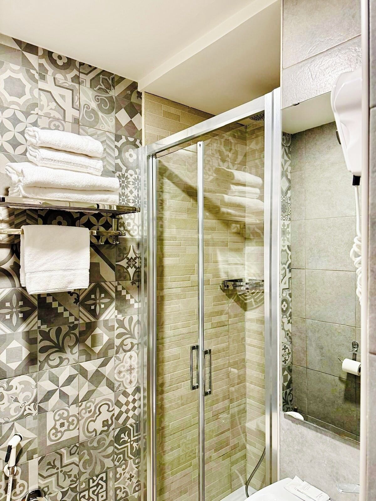 A modern bathroom with patterned tiles, a glass shower enclosure, and neatly folded towels on a shelf.