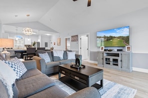 A modern living room with a large flat-screen TV, grey sofas, a coffee table, and a TV stand with shelves. - Serene Golf Retreat | Pool, Private Balcony, Grill (Myrtle Beach)