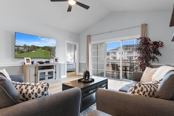 A spacious living room with a large flat-screen TV, a dining area with a table and chairs, and a balcony with a view of a golf course.