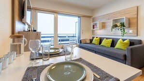 A modern living room with a dining area, large windows, a grey sofa with green cushions, and a mounted TV. - Remointze B 031 - CENTER studio 4 pers (Veysonnaz)