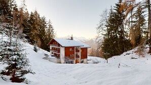 Een chalet met twee verdiepingen en een balkon, omringd door met sneeuw bedekte bomen en bergen op de achtergrond.