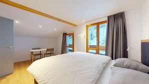 Ein Schlafzimmer mit einem großen Bett, einem Schreibtisch und Stühlen, einem Fenster mit Vorhängen sowie Blick auf Bäume.