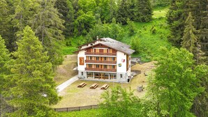 Un bâtiment de style chalet, entouré d’une forêt dense, avec un balcon bien visible et un chemin de pierre menant à l’entrée.