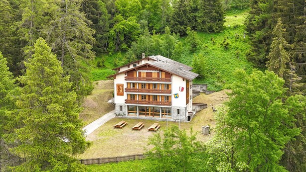 Ein Chalet-Stil-Gebäude, umgeben von dichtem Wald, mit sichtbarem Balkon und einem Steinweg, der zum Eingang führt.