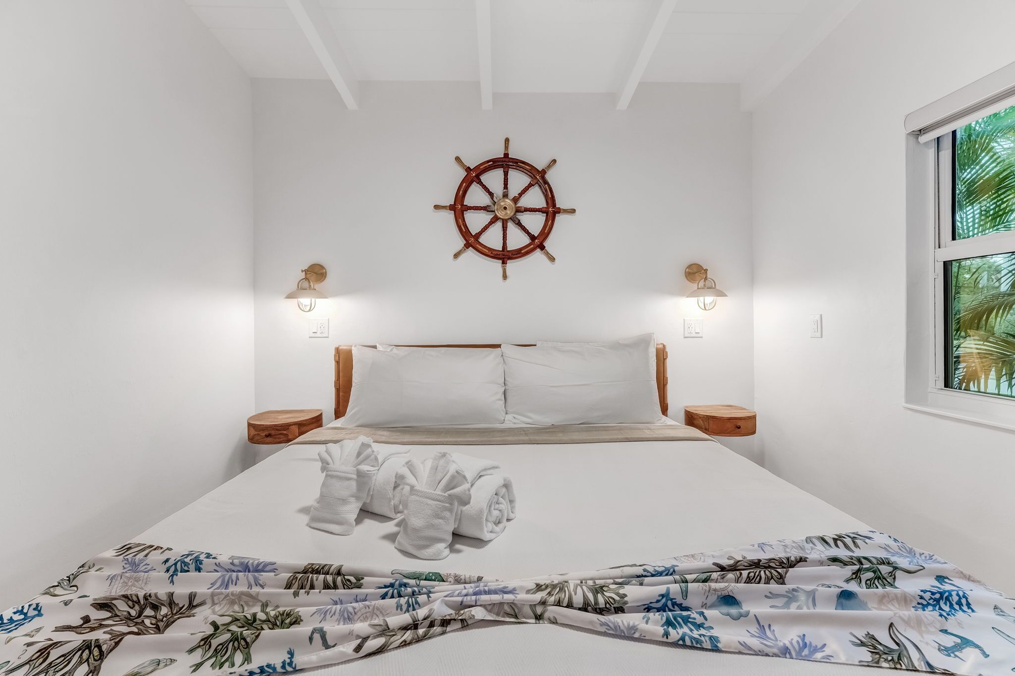 A neatly made bed with white linens, a ship's wheel on the wall, and a window with a view of palm trees.