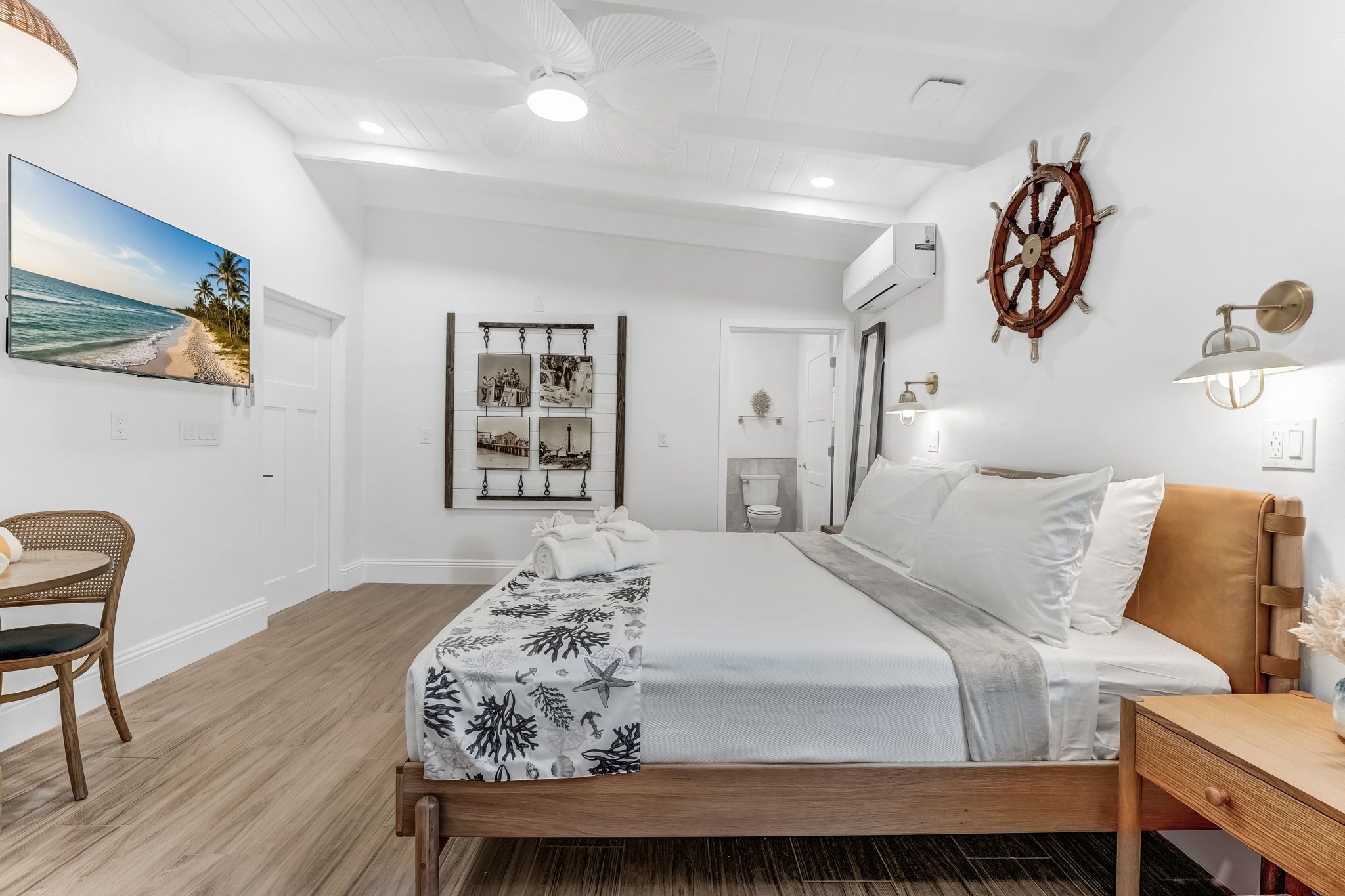 Un dormitorio con cama, escritorio, silla, ventilador de techo, una rueda de barco montada en la pared y un cuadro enmarcado con una playa.