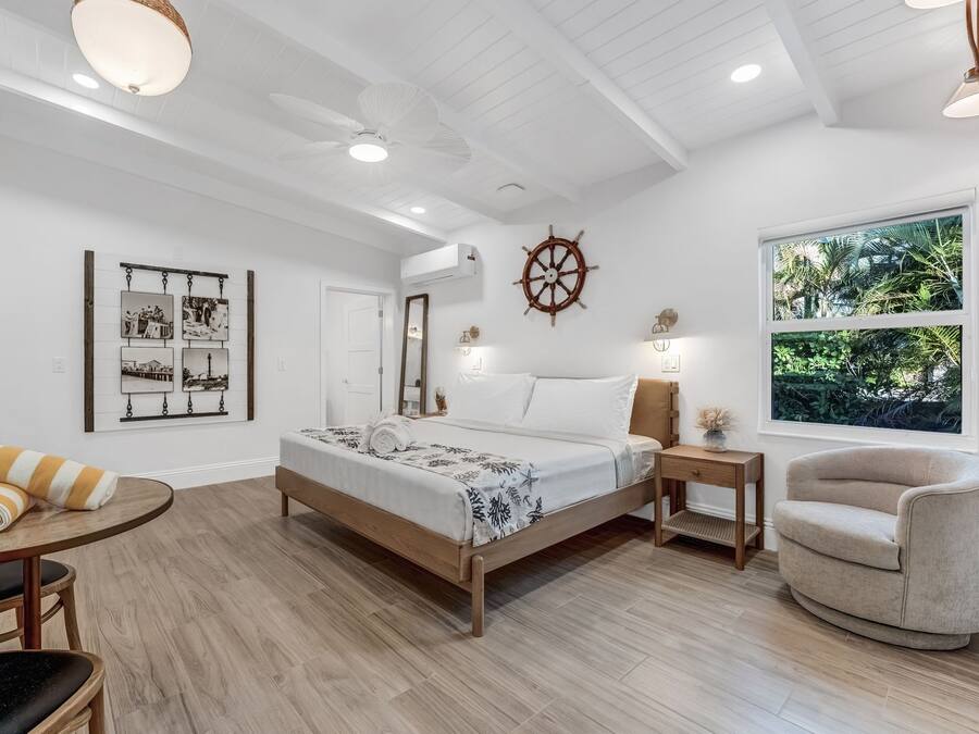 A bedroom with a bed, a round table, a chair, a TV, a ceiling fan, and a window with a view of greenery.