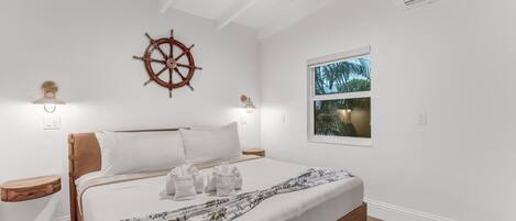 Un dormitorio con cama, un adorno de timón de barco, dos lámparas de noche, una ventana con vistas a palmeras y suelo de madera.
