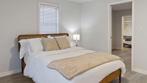 Un dormitorio con cama de madera, ropa de cama blanca, una manta beige, una lámpara de noche y una ventana con persianas.