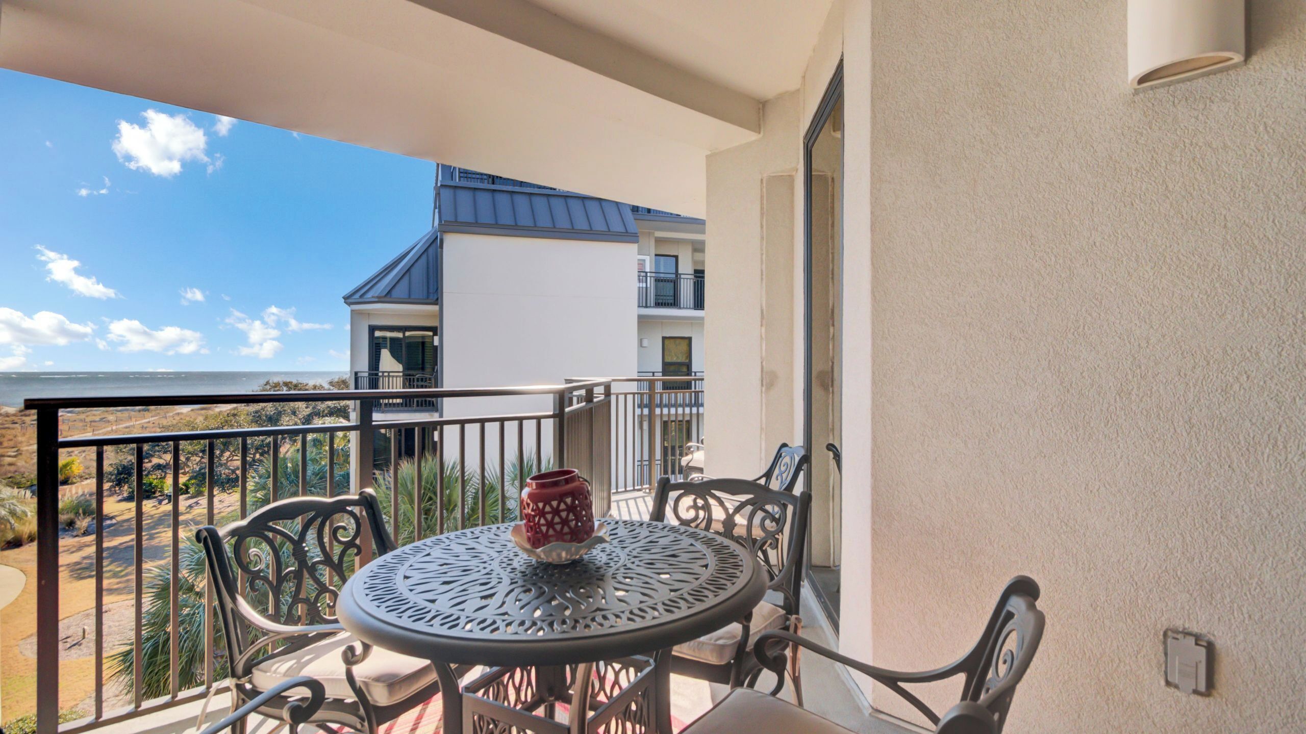 A balcony with a table and chairs, a red vase, and a view of a building and beach.