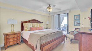 A hotel room with a wooden bed, two bedside tables with lamps, a ceiling fan, a balcony with a view, and a desk with a chair.