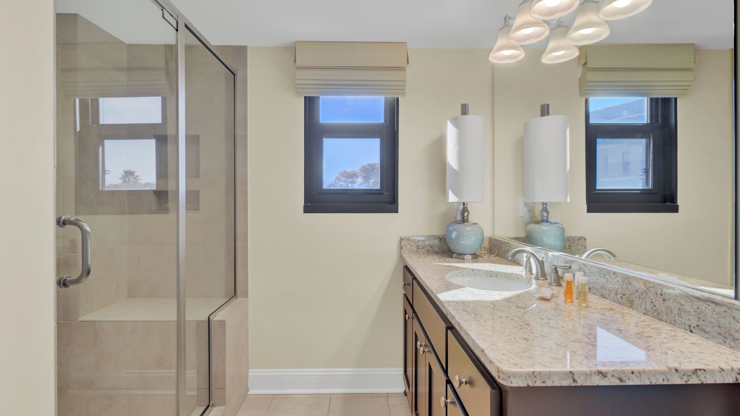 A bathroom with a glass shower enclosure, two windows with blinds, a marble countertop, and a mirror.
