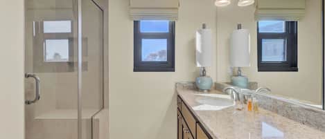 A bathroom with a glass shower enclosure, two windows with blinds, a marble countertop, and a mirror.