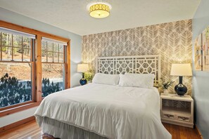 A bedroom with a large bed, two bedside tables with lamps, a decorative headboard, and a view of trees through large windows.