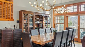 A dining area with a wooden table, leather chairs, a chandelier, and a large window with a view. - Mountain Escape With Game Room, Balcony & Stunning Views in the Poconos Christina (Tannersville)