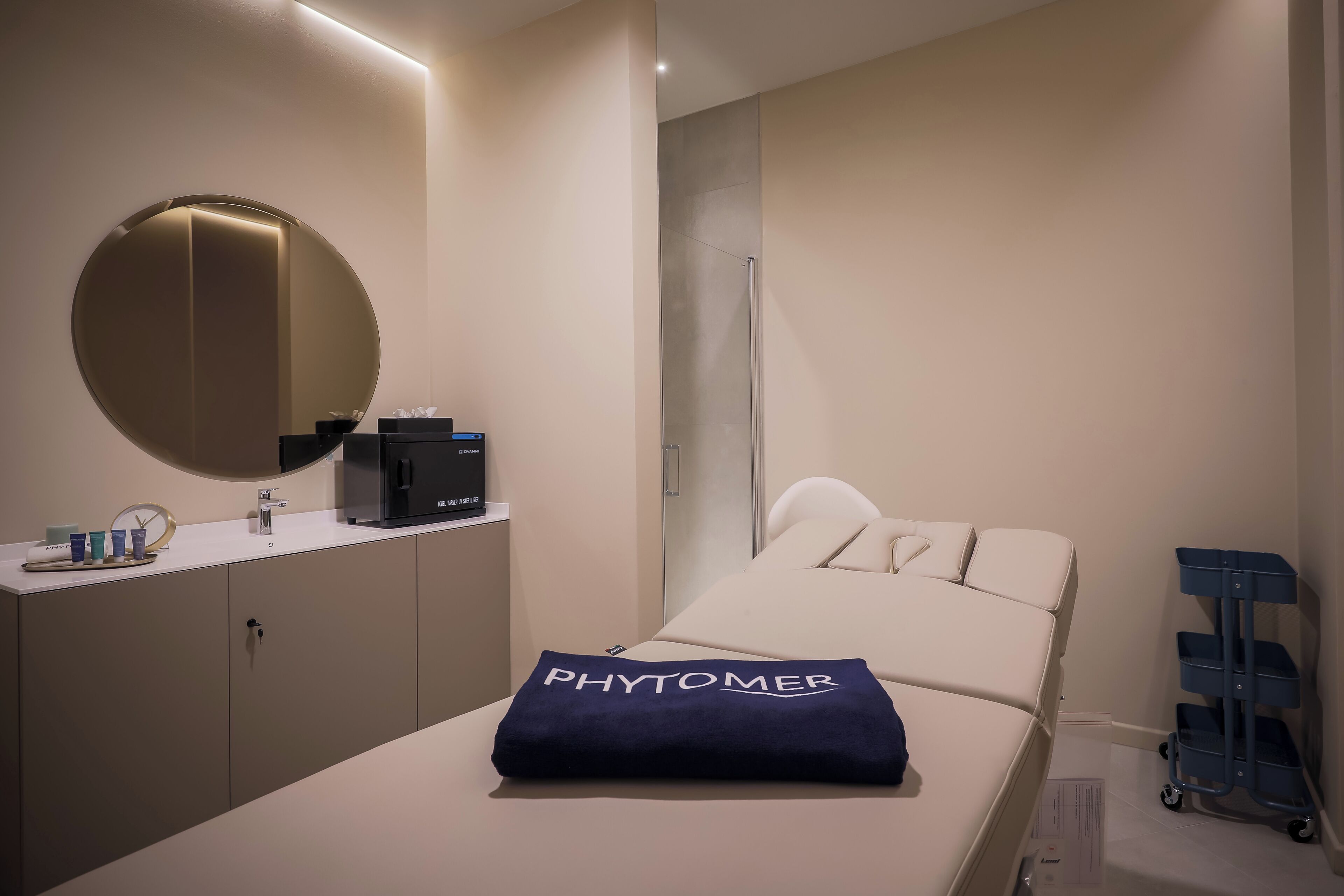 Ein modernes Hotelzimmer mit einem Bett, einem Nachttisch mit einer Kaffeemaschine, einem Spiegel und einem blauen Servicewagen.