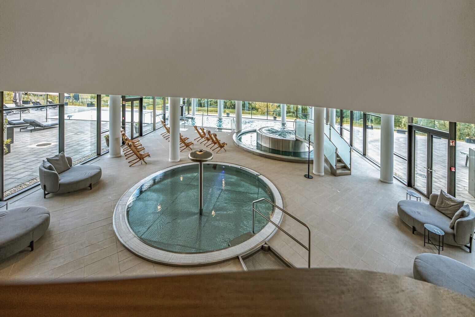 Et moderne poolområde indendørs med et centralt spabad, omgivet af liggestole og en trappe op til en etage over.