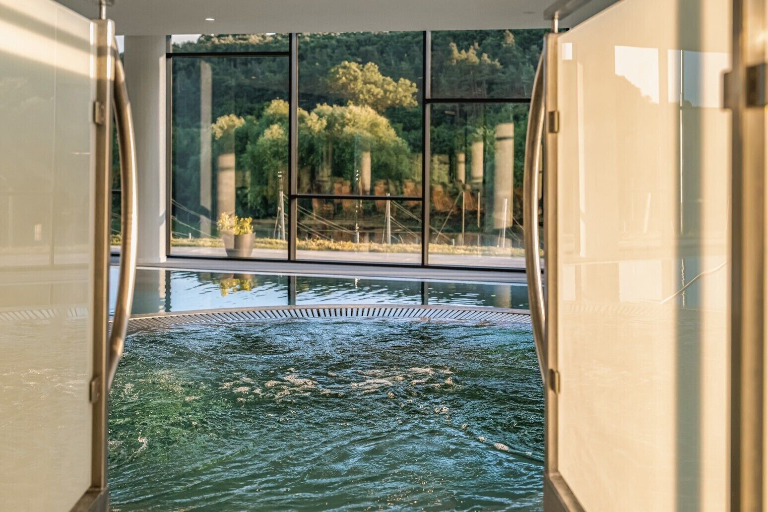 Une piscine intérieure moderne avec des parois en verre transparent, offrant une vue sur la verdure environnante et une surface d’eau calme.