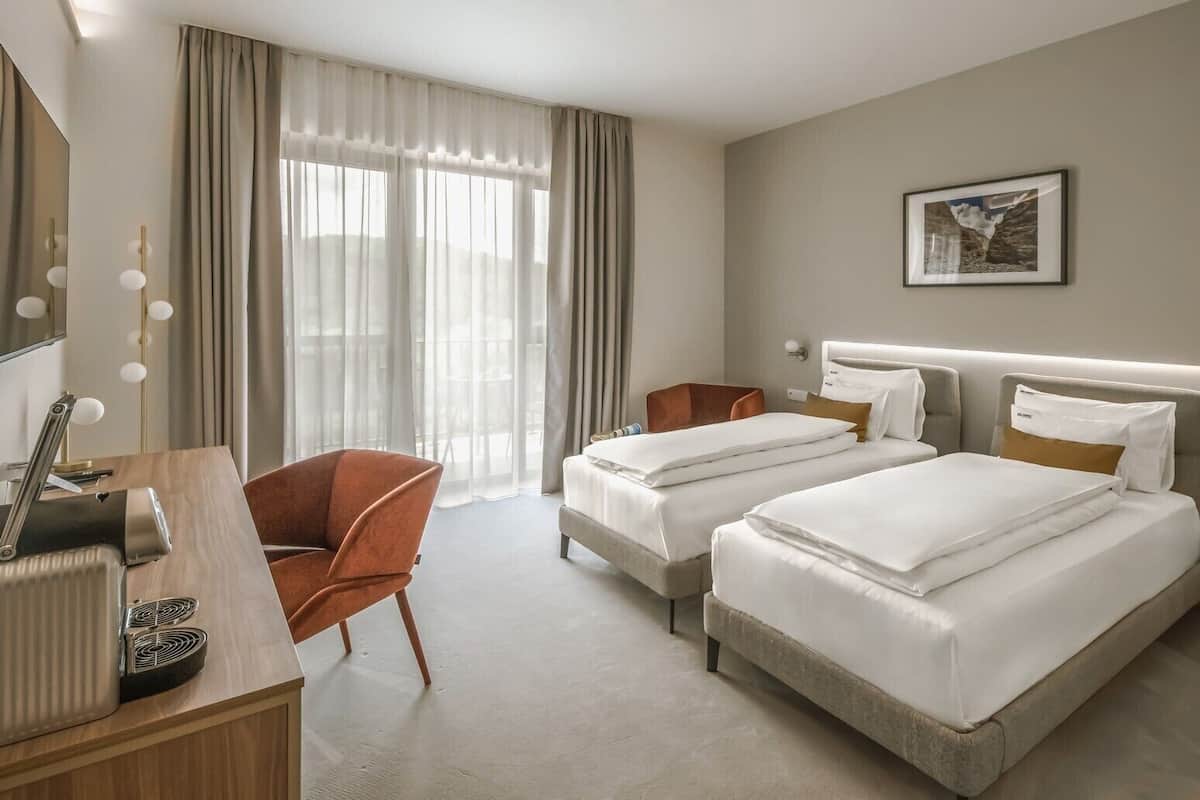Quarto de hotel com duas camas, uma mesa com abajur, uma cadeira e uma janela grande com cortinas.