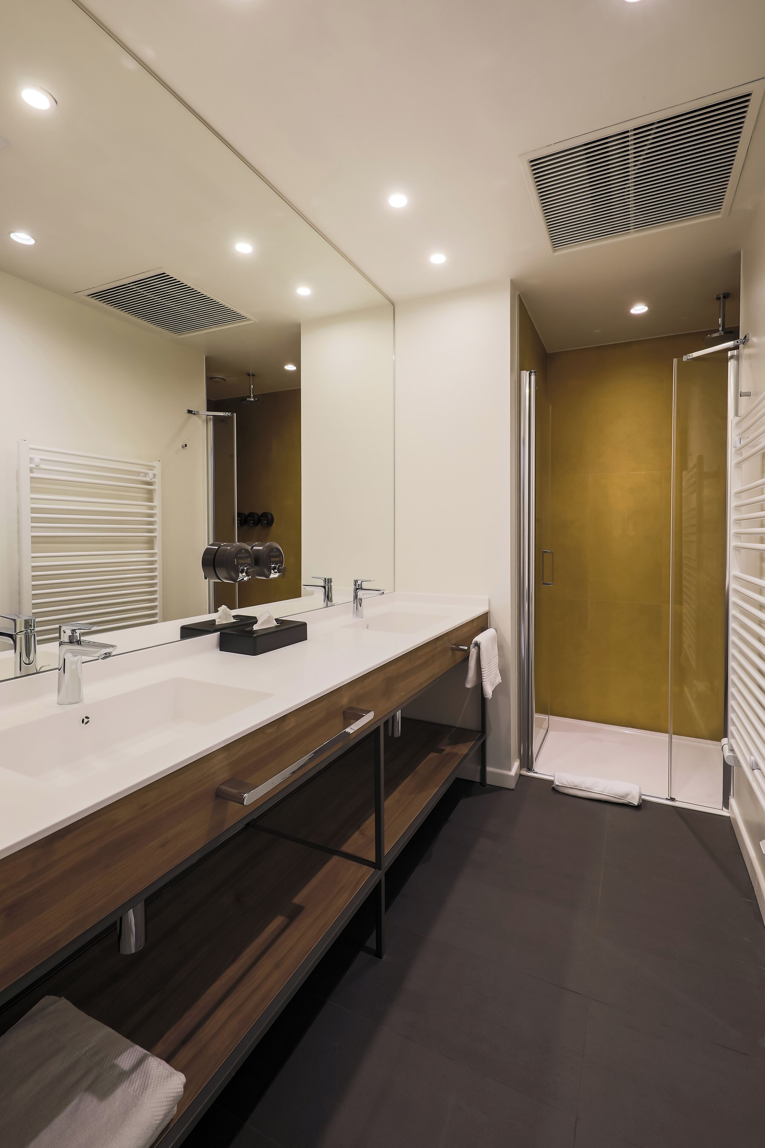 A modern bathroom with a large mirror, two sinks, a wooden vanity, a towel rack, and a shower with a gold door.