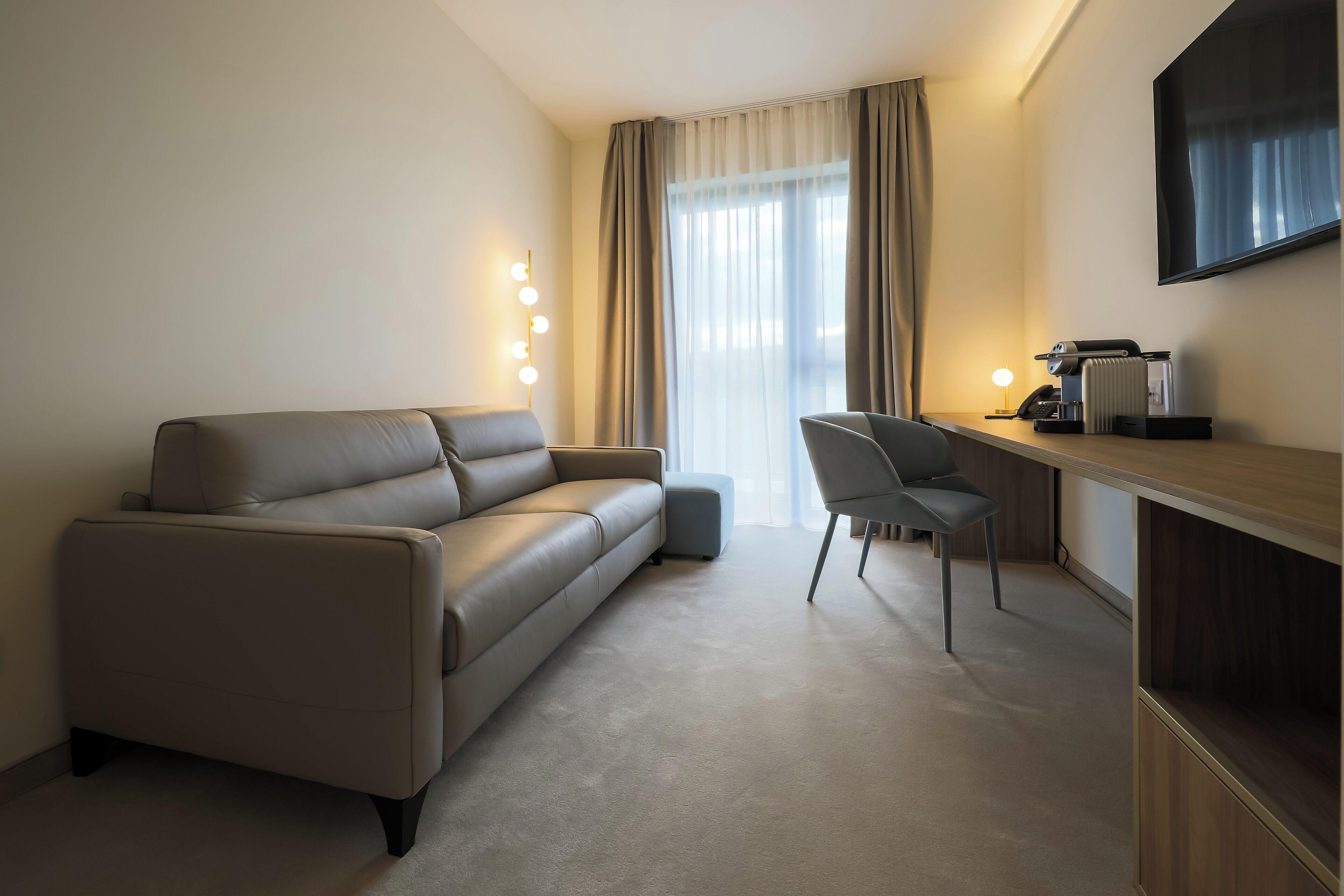 A modern hotel room with a sofa, a chair, a desk with a computer, and a TV mounted on the wall.