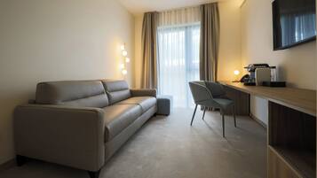 A modern hotel room with a sofa, a chair, a desk with a computer, and a TV mounted on the wall.