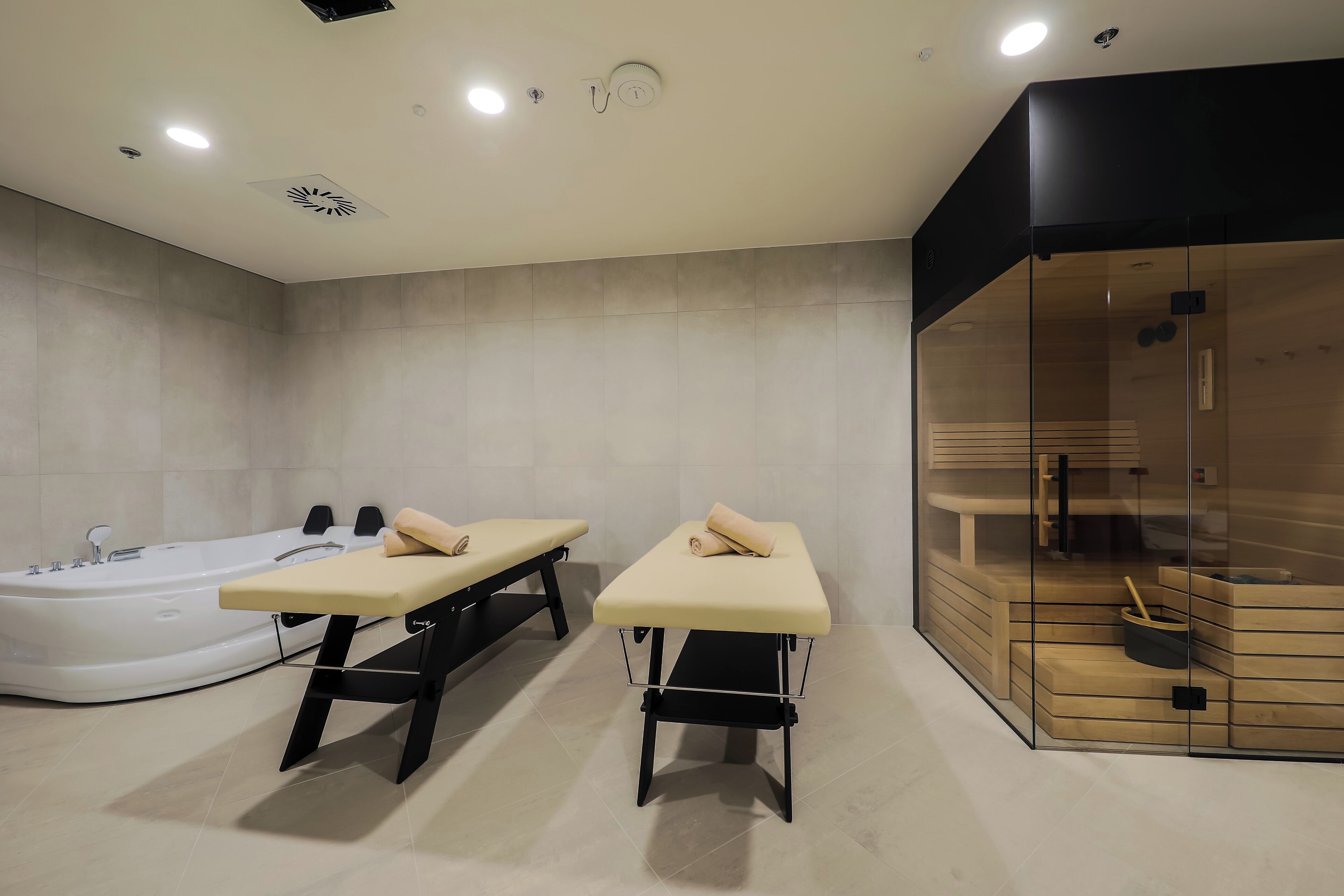 A modern spa room with a large white bathtub, two massage tables, and a glass-enclosed sauna area.