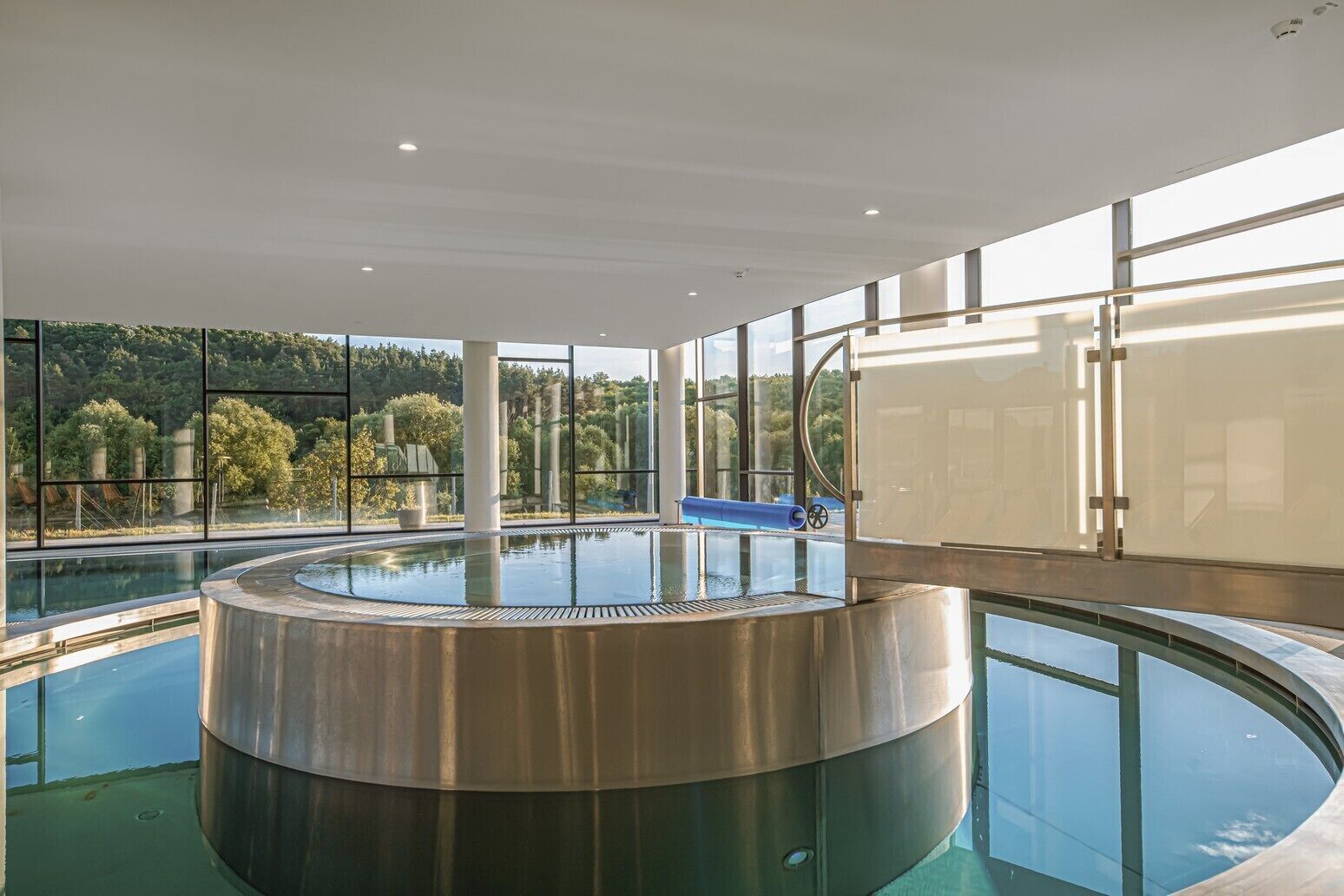 Ein moderner, innenliegender Schwimmteich mit kreisförmigem Design, großen Fenstern und Blick auf eine bewaldete Gegend sowie einer blauen Rutsche.