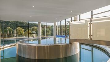 Ein moderner, innenliegender Schwimmteich mit kreisförmigem Design, großen Fenstern und Blick auf eine bewaldete Gegend sowie einer blauen Rutsche.