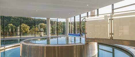 A modern indoor swimming pool with a circular design, large windows offering views of a forested area, and a blue slide.