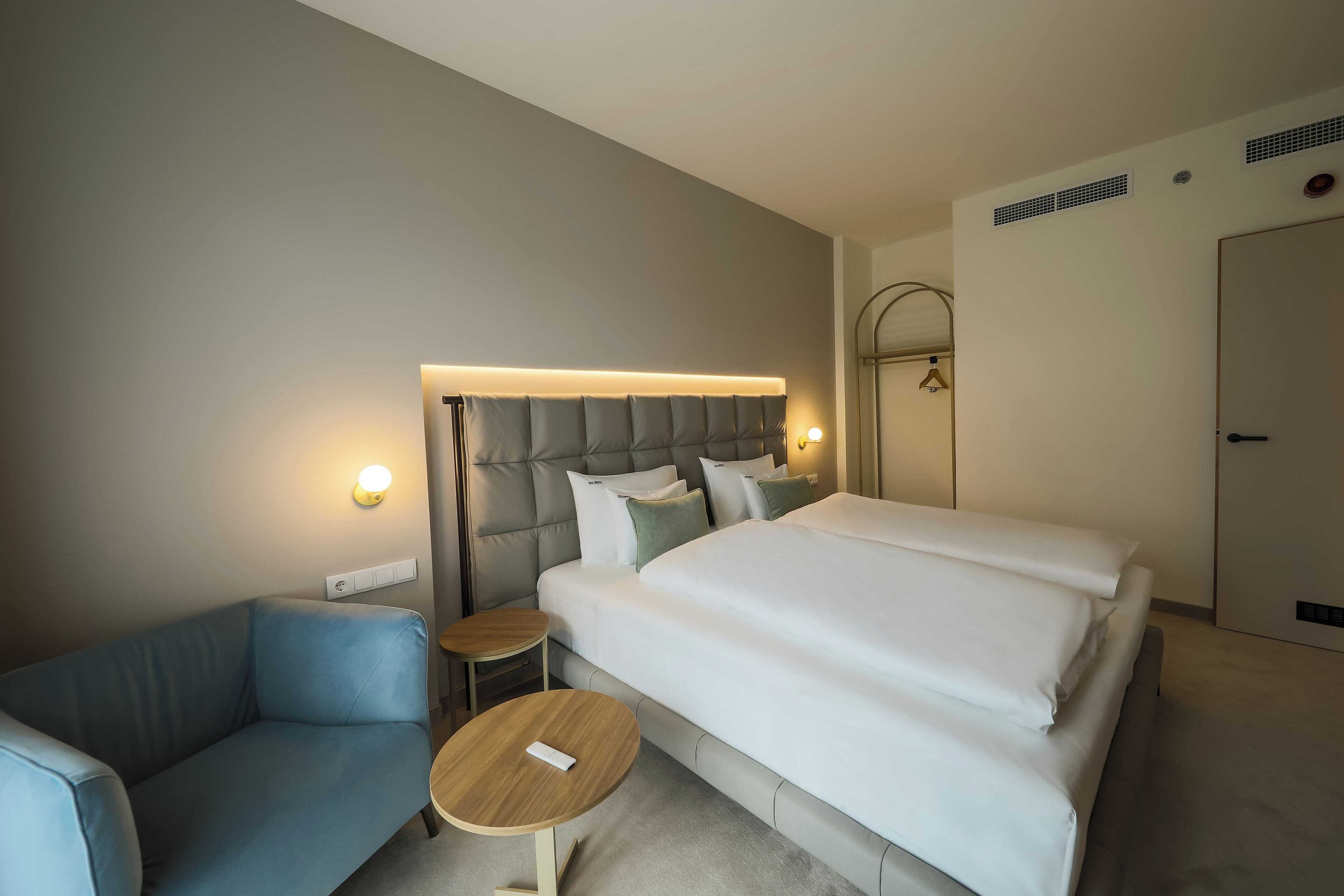 A modern hotel room with a large bed, a sofa, a small table, and a wall-mounted light fixture.