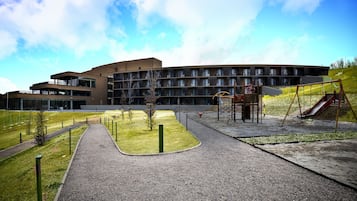 En modern byggnad med en lekplats, inklusive en rutschkana och gungor, omgiven av en grusgång och grönområden.
